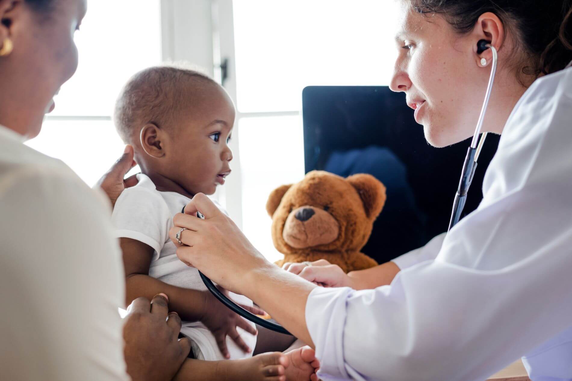 Doctor with stethoscope observing baby with mother nearby.