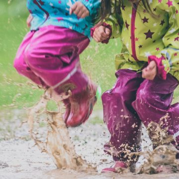 Two kids playing in a puddle with rain coats and waterproof pants.