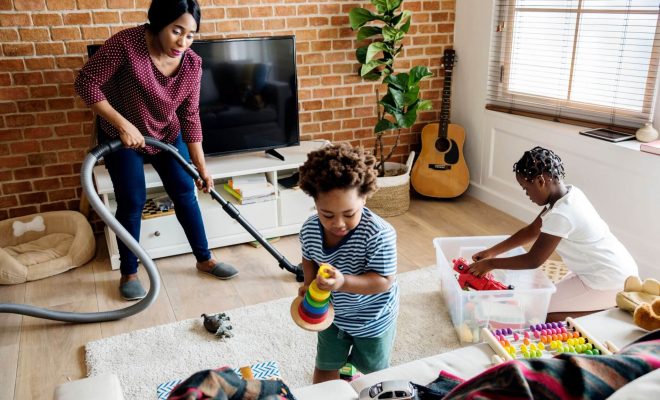 Family working together to clean a living room. The woman uses a vacuum, while the kids clean up toys.