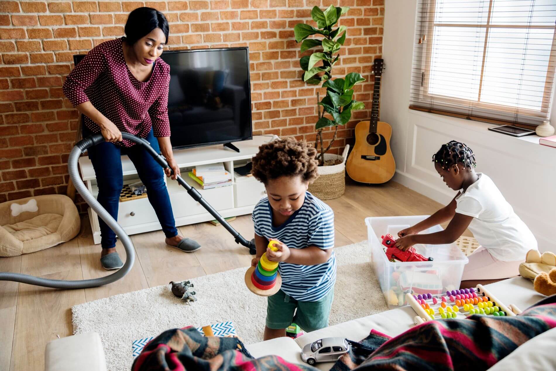 Family working together to clean a living room. The woman uses a vacuum, while the kids clean up toys.
